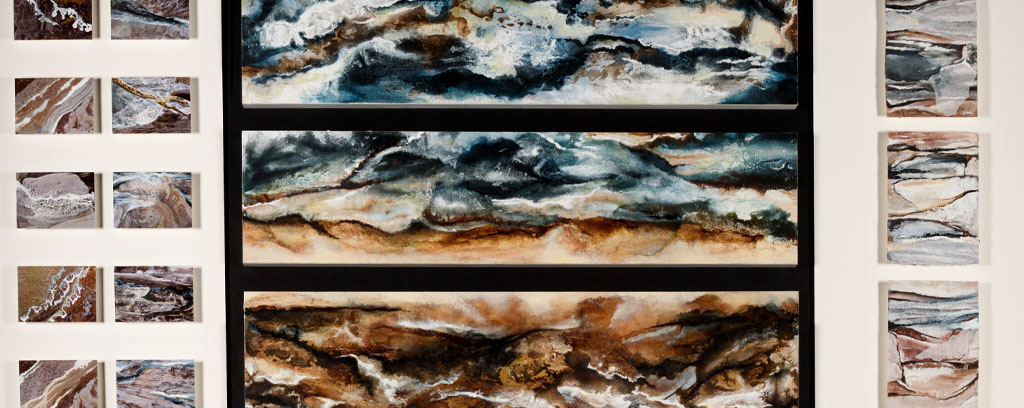 watercolour paintings of rough landscapes in earthy tones and blues, four long panels and thirteen smaller panels, close-ups of rocks and water.